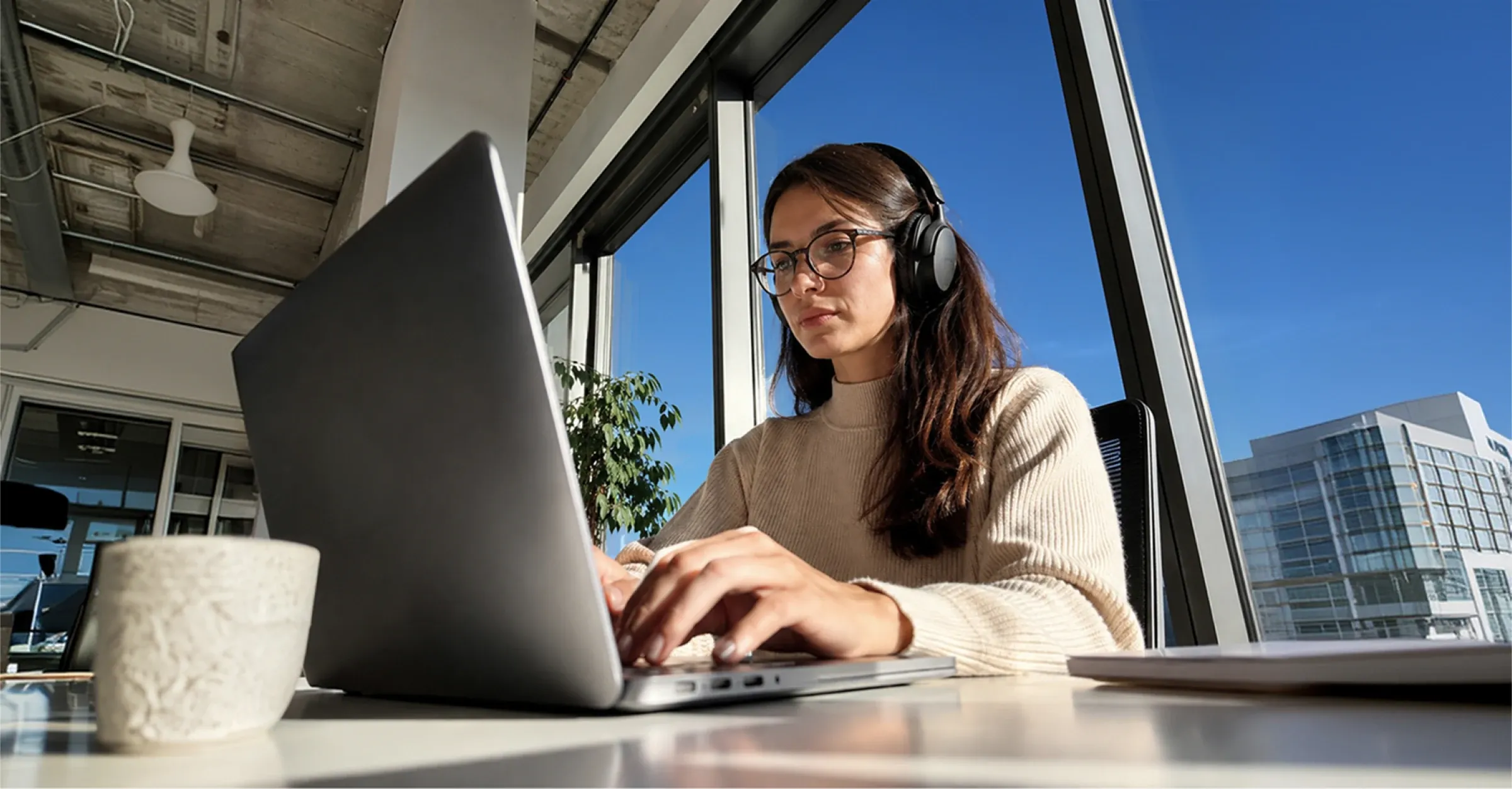 A woman with headphones works on a laptop near a window